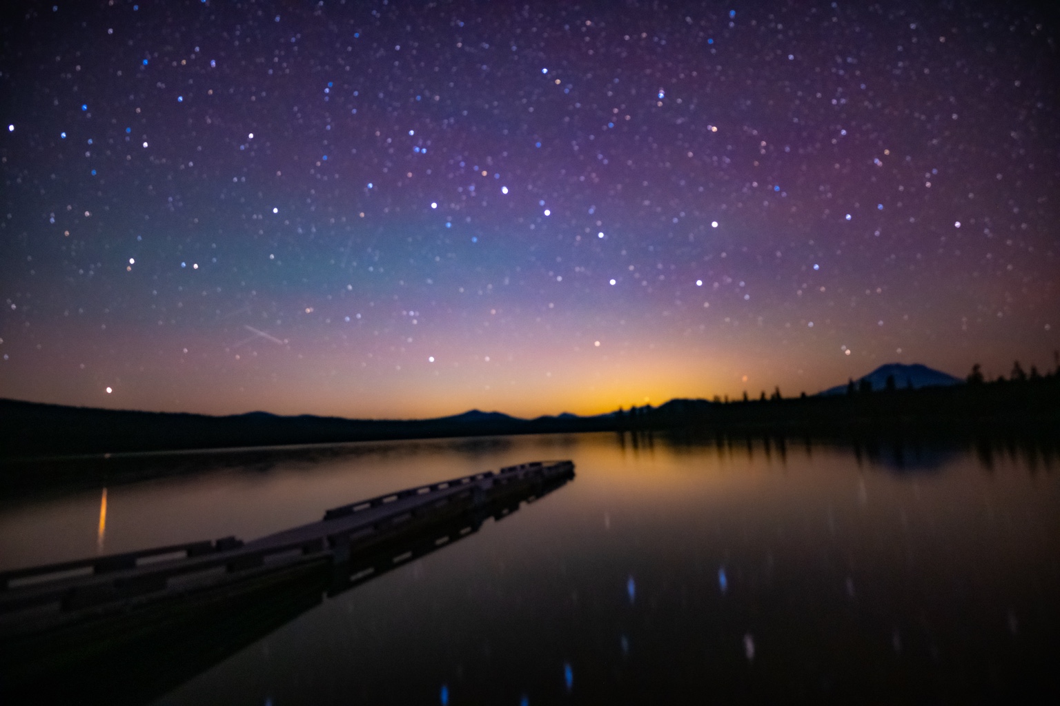 A wooden dock reaches into a still lake beneath a starry twilight sky