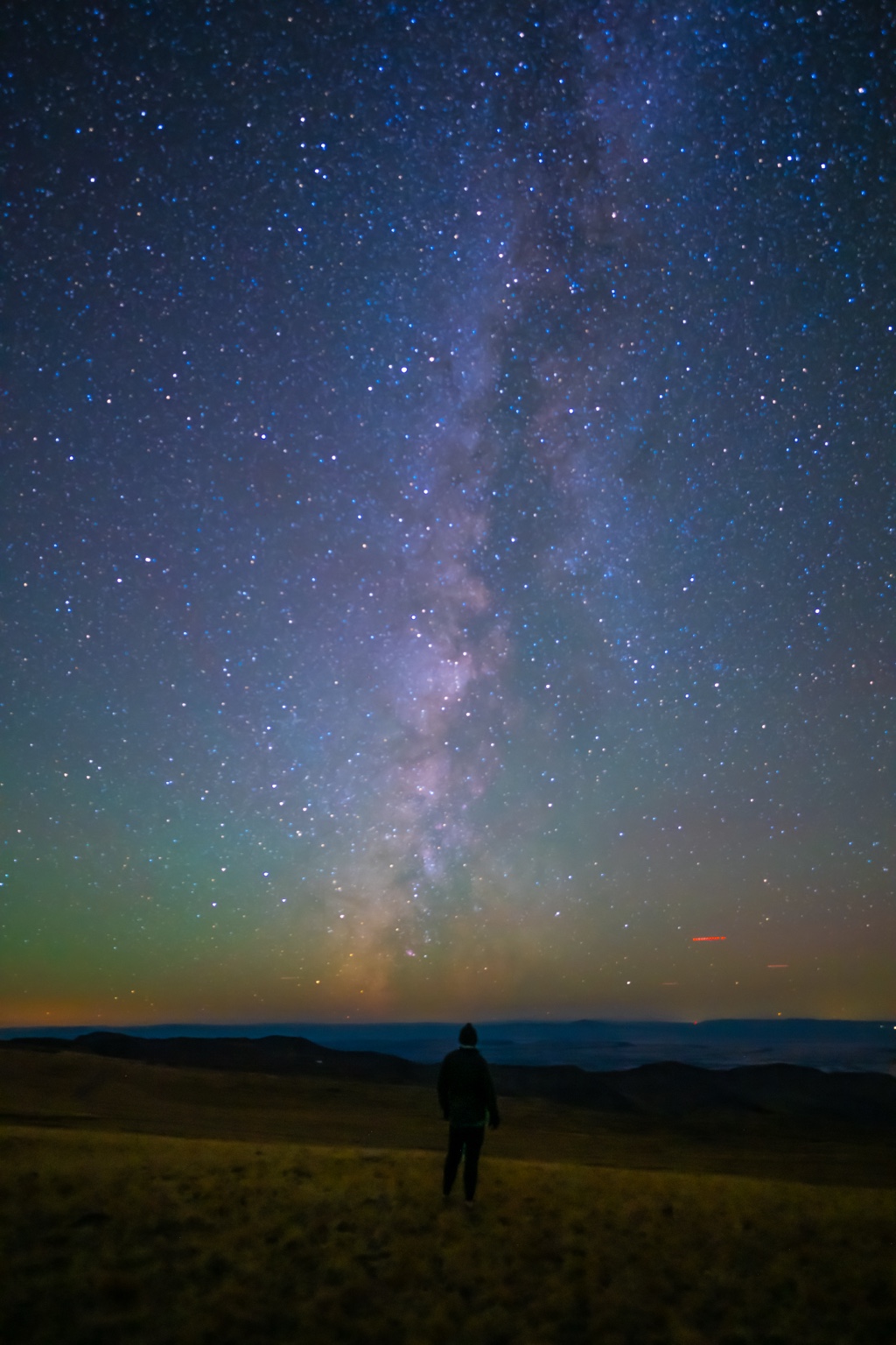 A lone figure beneath a vertical Milky Way and colorful horizon