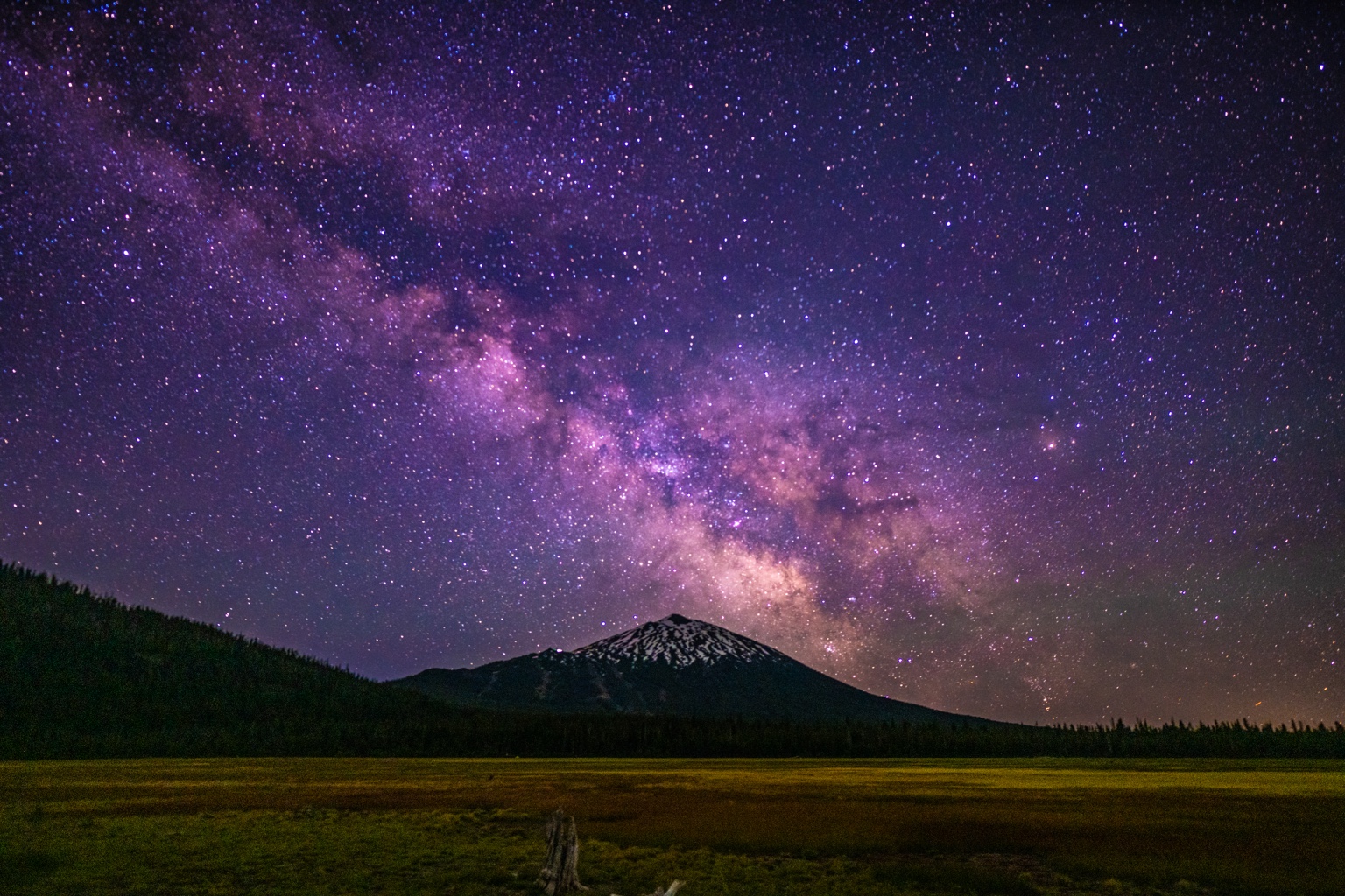 A purple Milky Way stretches above a snow-capped mountain and meadow